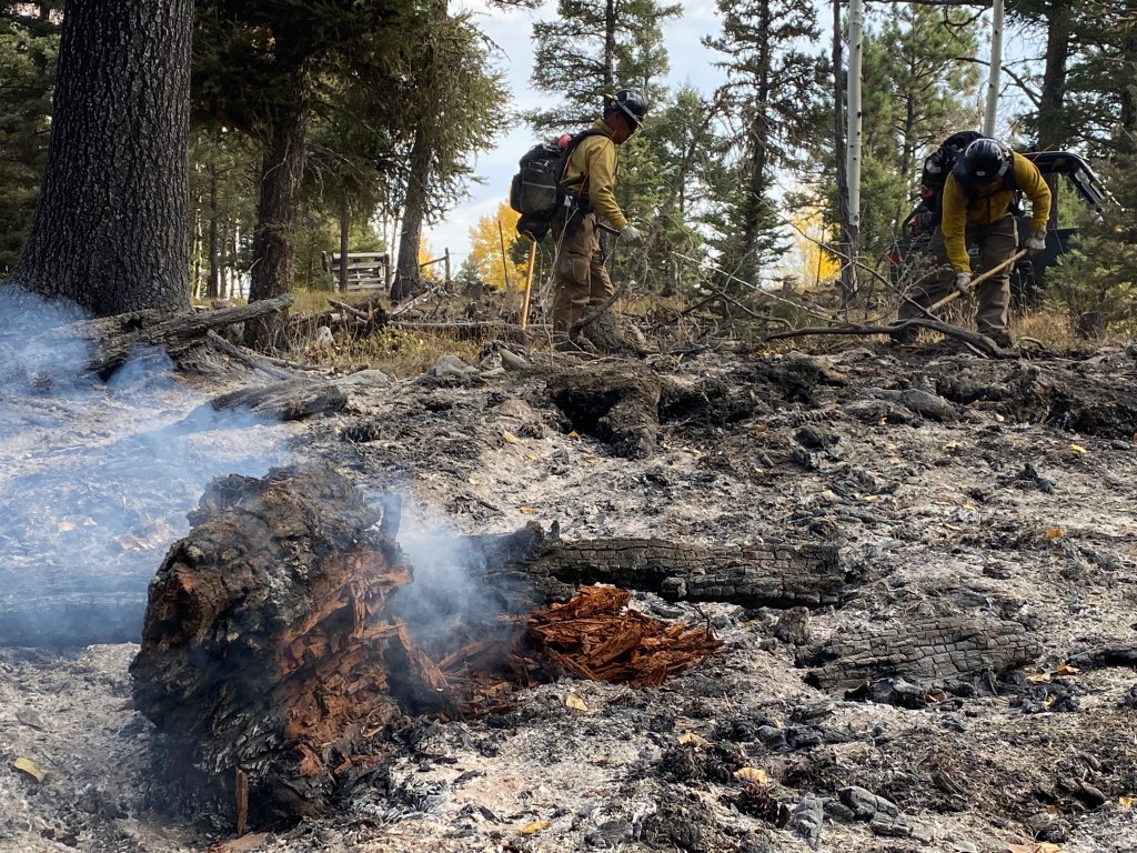Two firefighters mop up hotspots with water and a shovel near Taos Pines. A log smolders in the foreground.
