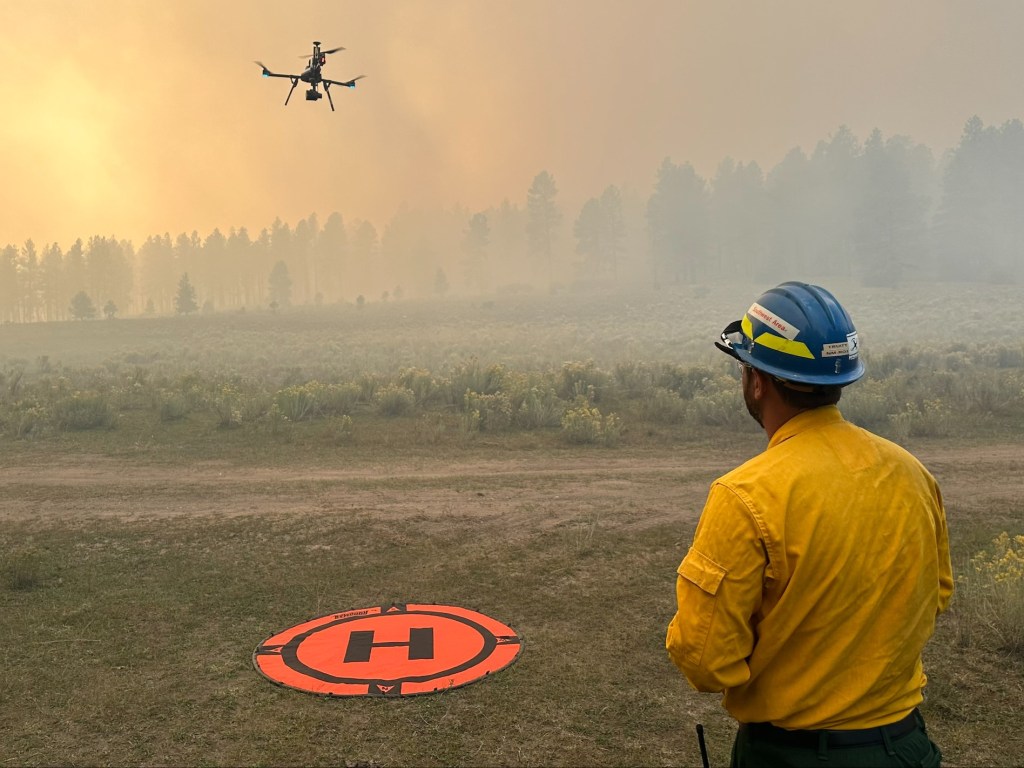 A fire crew member watches a drone lift off in a smoky forest.