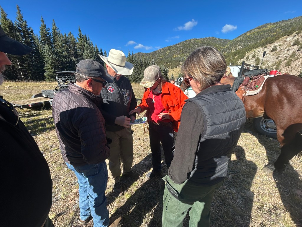 Four people in an open forest examine a piece of paper held by one of them.