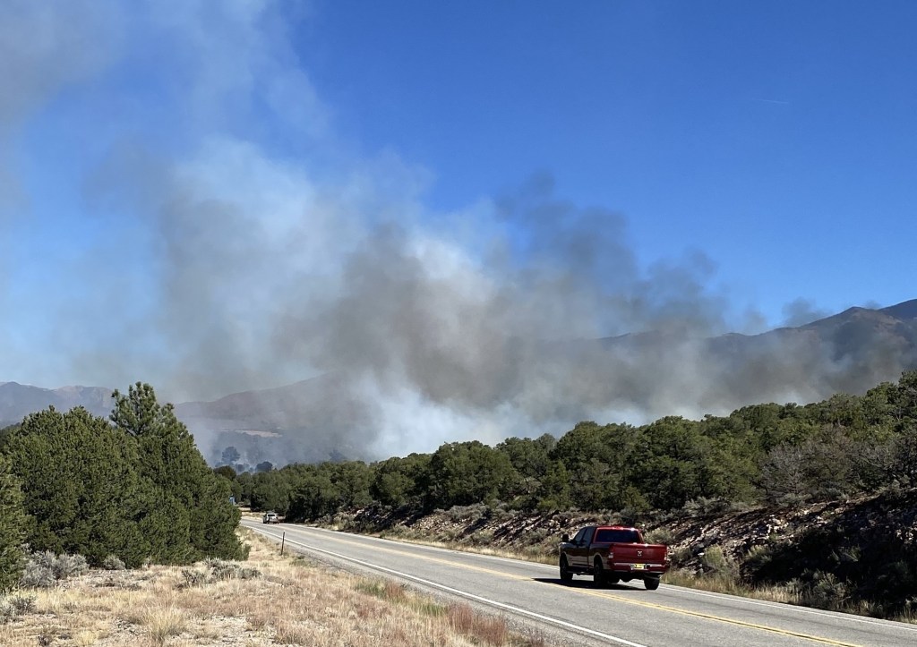 Smoke rises from a forest next to a paved two-land highway.