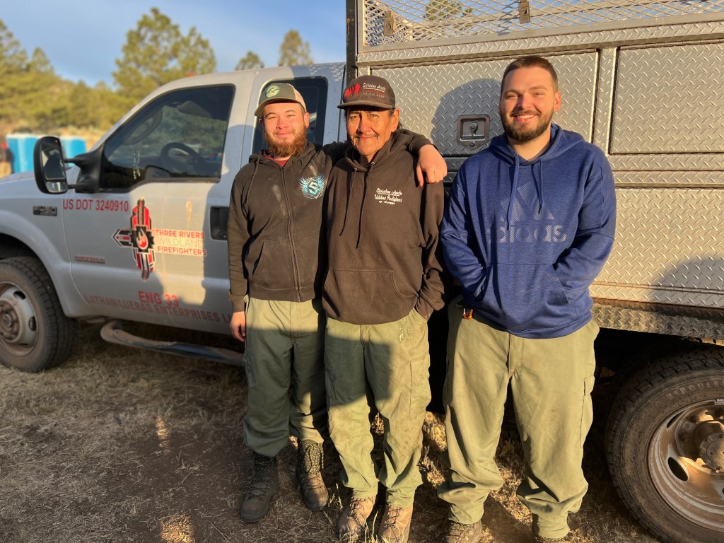 Three men stand in front of a wildland fire fighting brush truck adorned with a red and black zia symbol