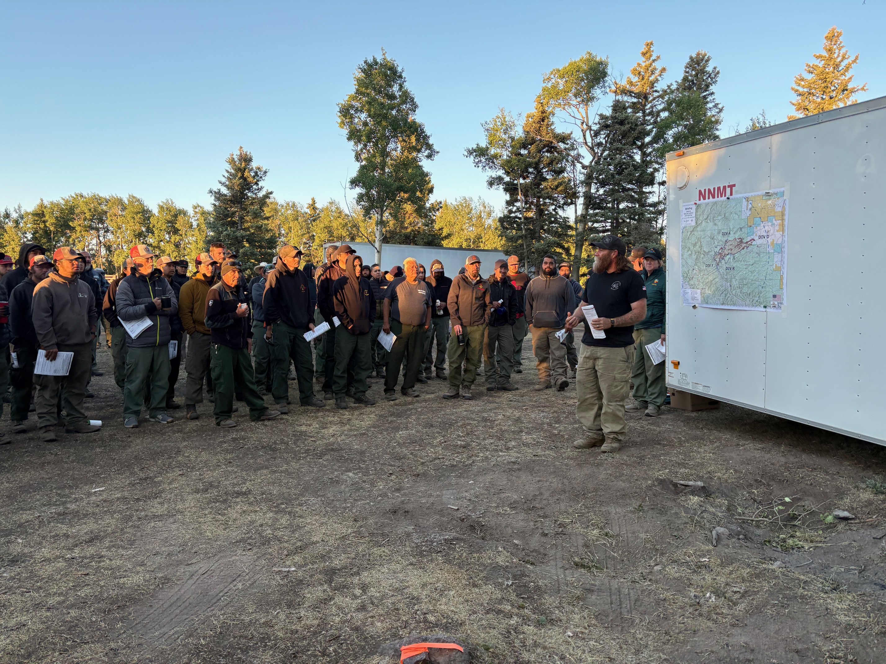 Incident Commander Dominic Sanchez stands in front of a map mounted on a trailer and speaks to a large group of firefighters during the morning briefing for the Beehive Fire on July 1, 2025, at the newly established operations base near Hopewell Lake. Trees and clear morning skies are visible in the background.