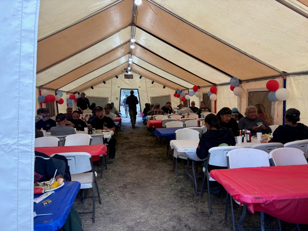 Inside the Incident Command Post (ICP) dining tent at the Beehive Fire, firefighters and support personnel sit at long tables decorated with red, white, and blue tablecloths and balloons, enjoying a Fourth of July dinner together. The large tent is filled with crews eating, talking, and taking a break from wildfire operations.